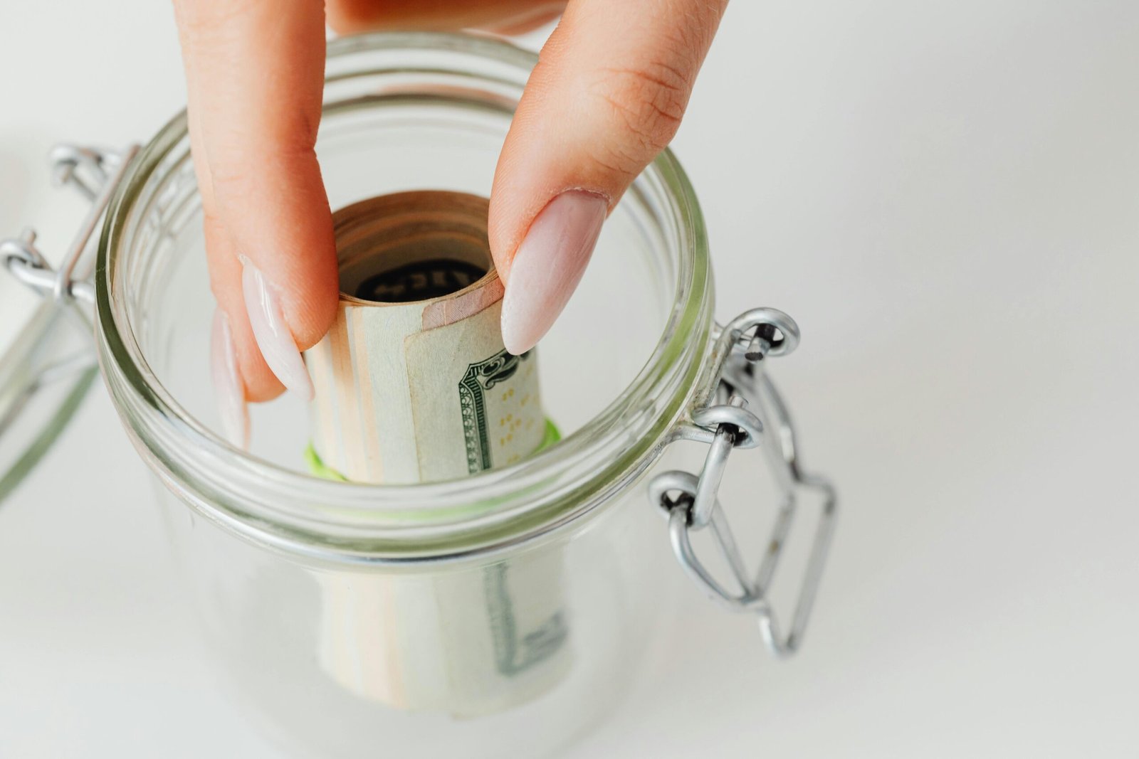 Close-up of hand placing rolled currency into a clear glass jar, symbolizing savings.