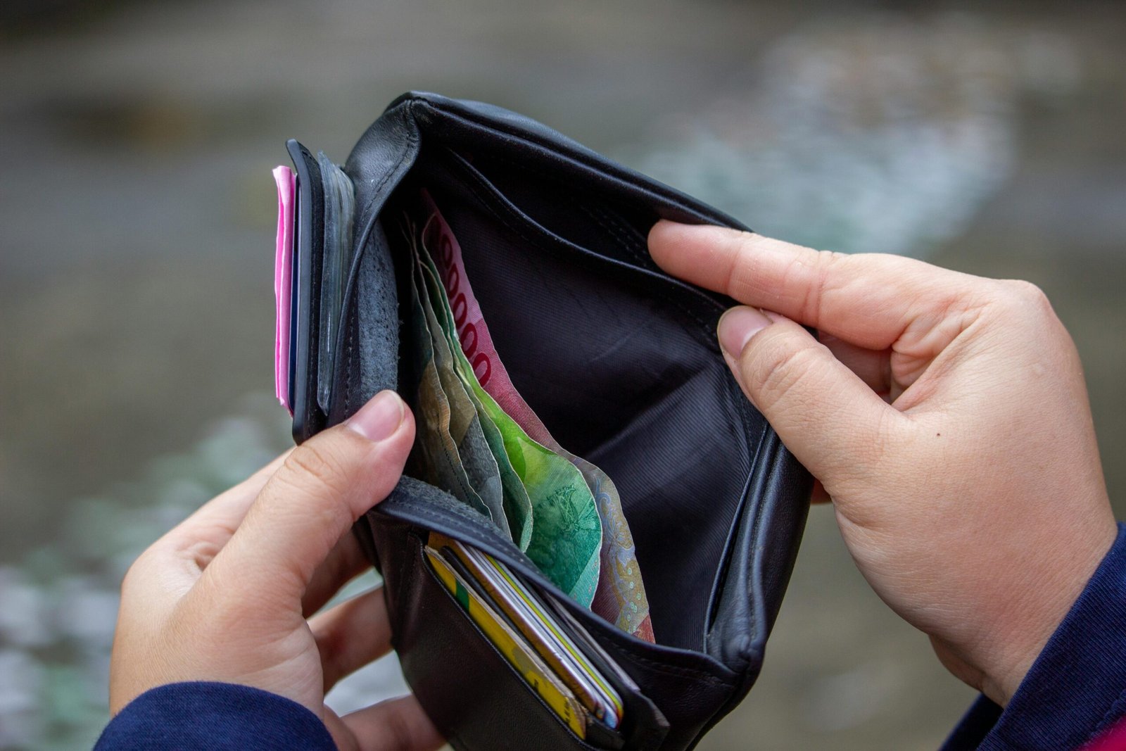 Person holding open wallet revealing banknotes, including Indonesian rupiah, in an outdoor setting.